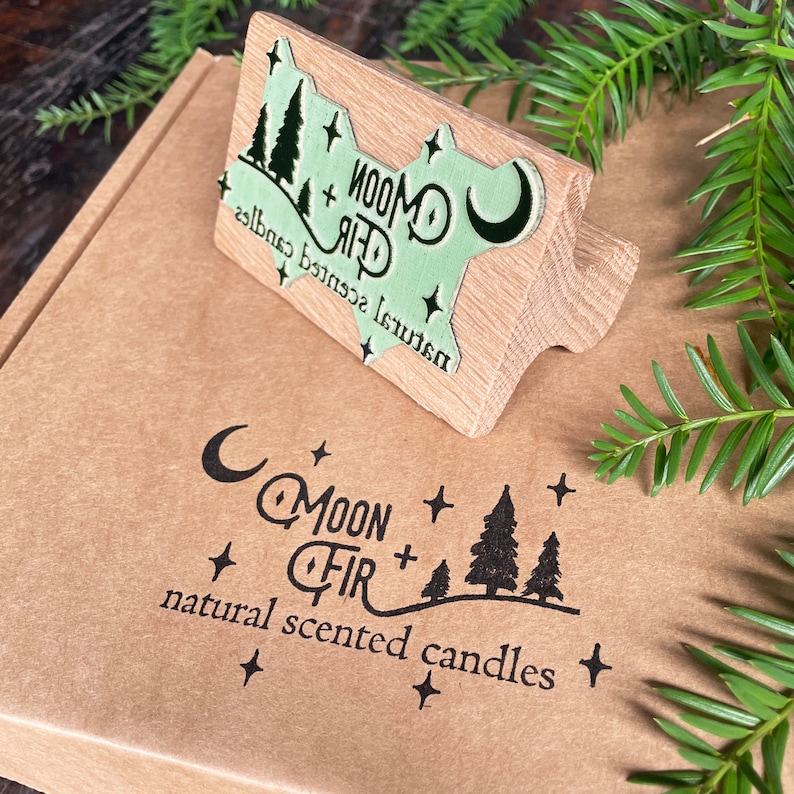 Pu&ograve; includere: Un timbro di legno con un design verde e bianco che dice "Moon + Fir Natural Scented Candles". Il timbro &egrave; sopra una scatola marrone con lo stesso logo stampato in nero.