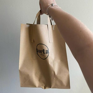 Pu&ograve; includere: Un sacchetto di carta marrone con un logo bianco che dice "torta bakery" in lettere nere.