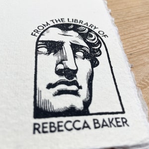 May include: Black and white stamp with a bust of a person's face in an archway. The text "From the Library of" is above the bust and "Rebecca Baker" is below the bust.