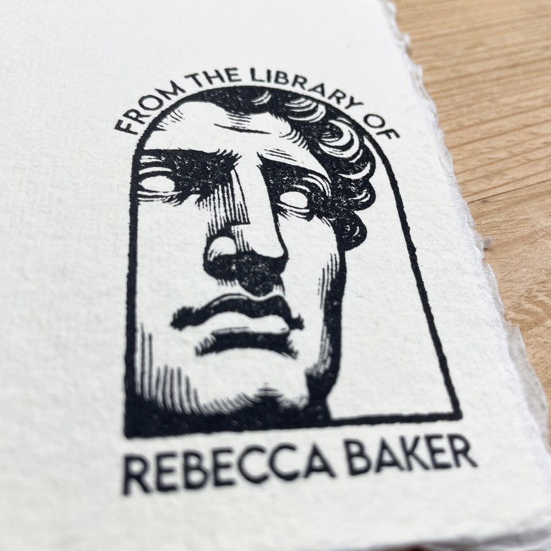 May include: Black and white stamp with a bust of a person's face in an archway. The text "From the Library of" is above the bust and "Rebecca Baker" is below the bust.