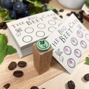 Pu&ograve; includere: Un timbro in legno con un design di barbabietola verde e nero. Il timbro &egrave; su un blocco di legno e si trova accanto a un mazzo di biglietti da visita bianchi che recitano "The Big Beet Cafe".