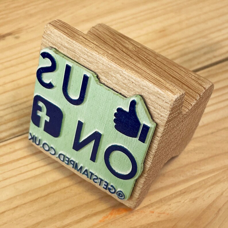 May include: A wooden stamp with a green and blue design. The stamp features the text "GET STAMPED CO. UK" and a thumbs up icon. The stamp is made of natural wood and has a rustic look.