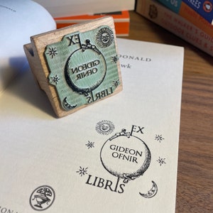 May include: A wooden stamp with a green and black design. The stamp features a globe with the text "Gideon Ofnir" and "Libris" around it. There are stars and a sun on the stamp.