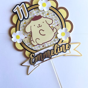 Puede incluir: Adorno para tarta de varias capas con un personaje de perro de dibujos animados, margaritas y el número 11. El adorno tiene una pancarta con el nombre "Emmeline" en dorado y negro. El diseño incluye elementos amarillos, marrones y blancos.