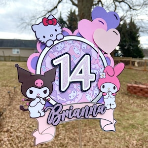 Puede incluir: Un adorno para pastel rosa y morado con los personajes Hello Kitty, My Melody y Kuromi. El adorno tiene un círculo morado con el número 14 y el nombre Brianna en letras moradas brillantes.