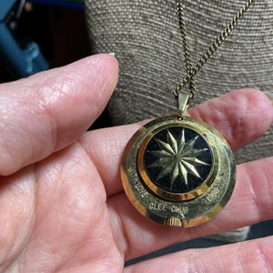 May include: A gold-toned pendant necklace with a black center featuring a starburst design. The pendant is round and has the words "WITH LOVE GLEE CLUB 1960" engraved around the edge. The chain is a delicate, matching gold color.