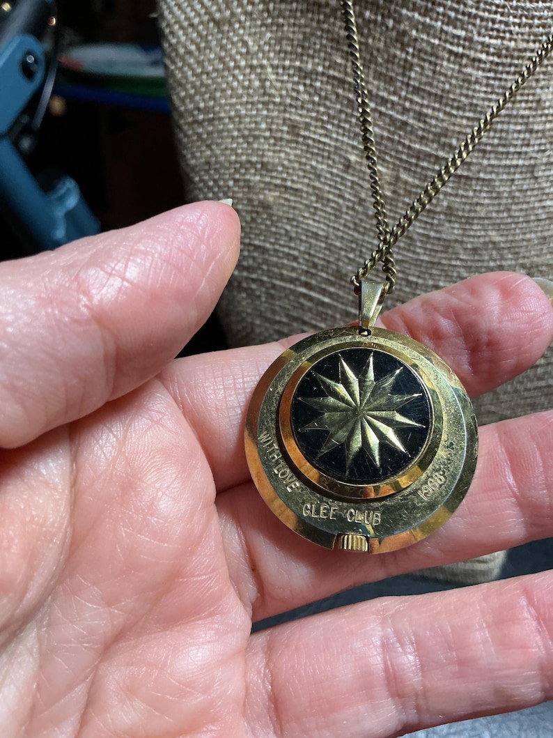 May include: A gold-toned pendant necklace with a black center featuring a starburst design. The pendant is round and has the words "WITH LOVE GLEE CLUB 1960" engraved around the edge. The chain is a delicate, matching gold color.