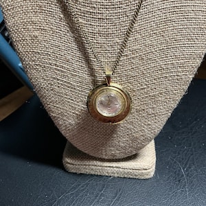 May include: A gold-toned pendant watch necklace. The round watch face has a light-colored dial with black numerals and hands. The watch is suspended from a delicate chain and displayed on a tan fabric bust.
