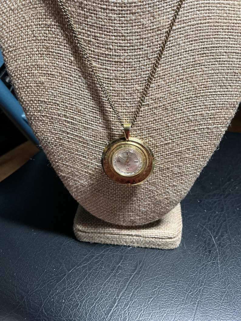 May include: A gold-toned pendant watch necklace. The round watch face has a light-colored dial with black numerals and hands. The watch is suspended from a delicate chain and displayed on a tan fabric bust.