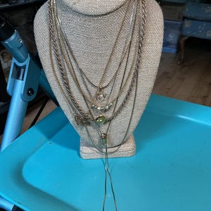 May include: A collection of layered gold-tone necklaces displayed on a beige fabric bust. The set includes various chain styles, a small heart pendant, a row of beads, a butterfly charm, a circular green stone pendant, and a long lariat-style chain.