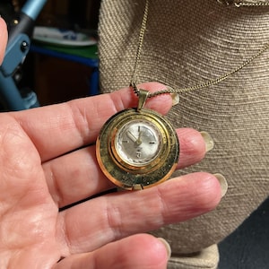 May include: A vintage gold-toned pendant watch with a round face and a delicate chain. The watch face has a silver-colored dial with black numerals and hands. The watch is displayed on a neutral-colored surface.
