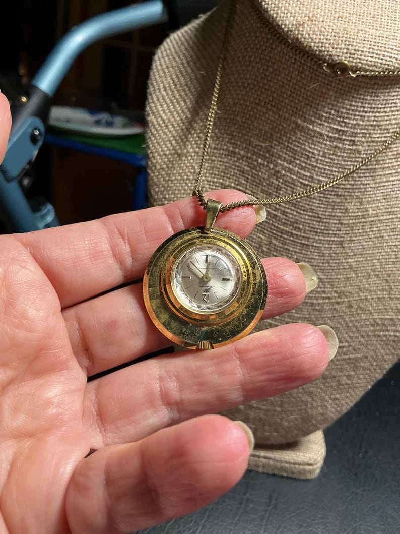 May include: A vintage gold-toned pendant watch with a round face and a delicate chain. The watch face has a silver-colored dial with black numerals and hands. The watch is displayed on a neutral-colored surface.