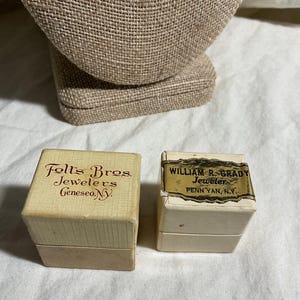 May include: Two small, vintage jewelry boxes. One box is beige with the text "Folts Bros. Jewelers Geneseo, NY." The other box is white with a label that reads "William R. Grady Jeweler Penn Yan, N.Y."