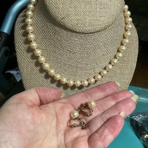 May include: A single-strand necklace featuring round, cream-colored faux pearls of uniform size, paired with a matching set of two screw-back earrings, each adorned with a single cream-colored faux pearl set in a gold-toned metal frame.