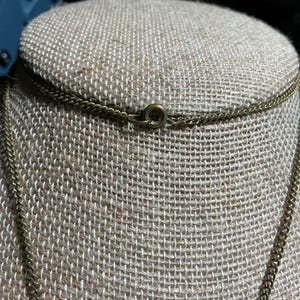 May include: Close-up of a gold-colored chain necklace with a small clasp. The necklace is displayed on a tan fabric surface. The chain has a delicate, textured appearance.