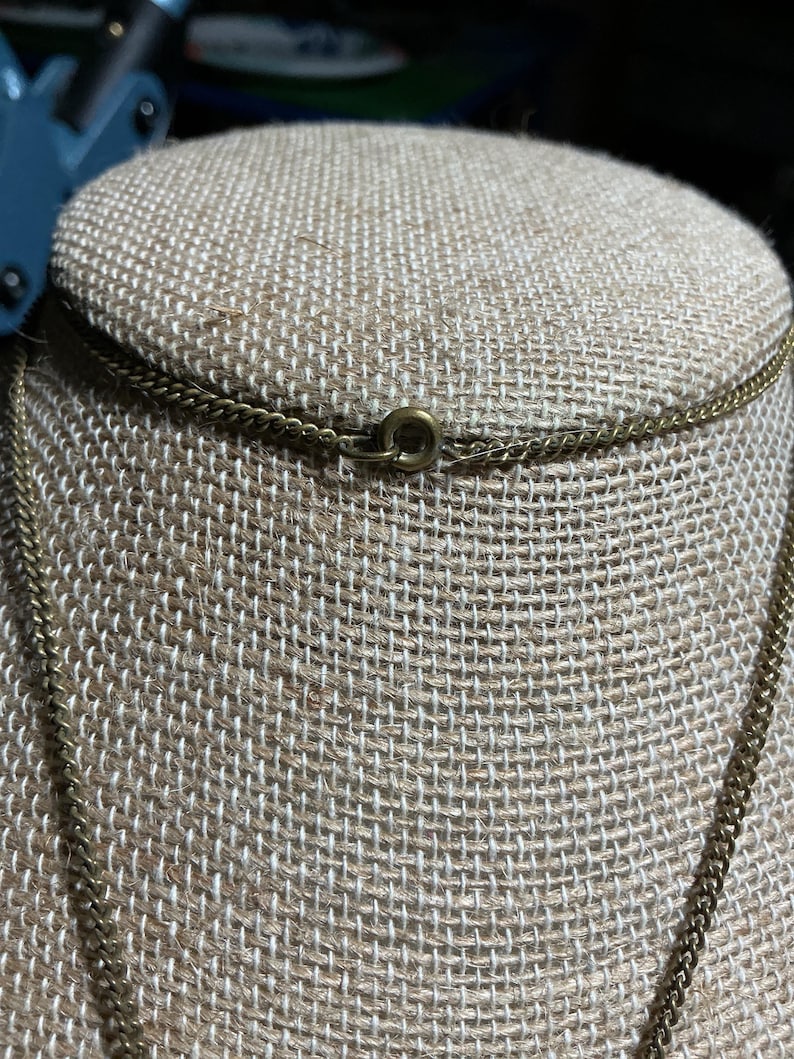 May include: Close-up of a gold-colored chain necklace with a small clasp. The necklace is displayed on a tan fabric surface. The chain has a delicate, textured appearance.