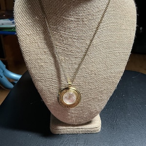 May include: A gold-toned necklace with a circular pendant. The pendant features a watch face design with a light-colored background and gold-colored hands and numerals. The necklace is displayed on a beige mannequin.