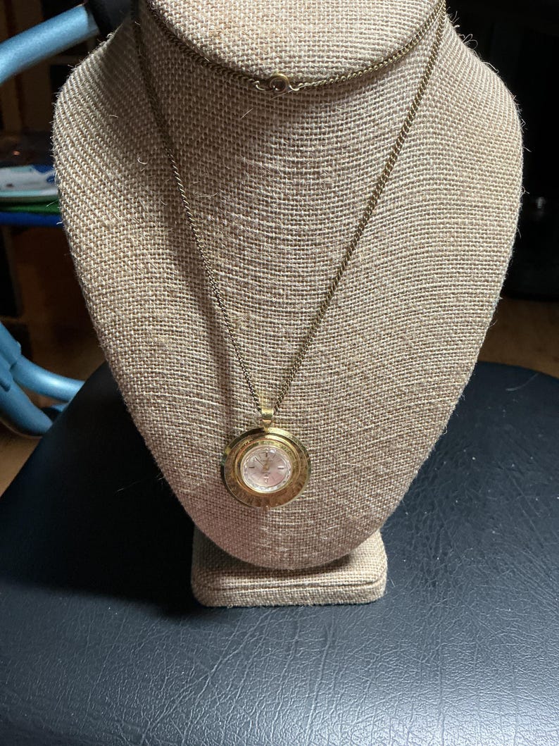 May include: A gold-toned necklace with a circular pendant. The pendant features a watch face design with a light-colored background and gold-colored hands and numerals. The necklace is displayed on a beige mannequin.