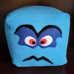 Op de afbeelding: Een blauwe pluche kubus met een gestikt gezicht. Het gezicht heeft zwarte wenkbrauwen, blauwe ogen met rode pupillen en een zwarte fronsende mond.