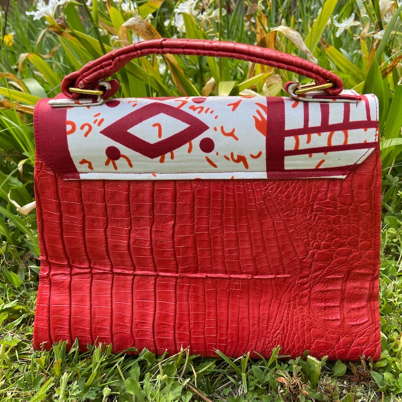 May include: A red crocodile-embossed leather handbag with a white and red patterned flap. The bag has a top handle and a gold clasp.