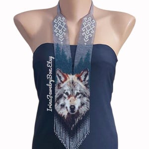 May include: A beaded necklace with a wolf design. The necklace features a geometric pattern in grey and white at the top, leading to a forest scene and a wolf's face. The bottom of the necklace has a white beaded fringe.