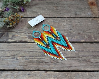 May include: A pair of long, beaded earrings with a turquoise, black, white, red, and yellow color scheme. The earrings are hanging from silver hoops.