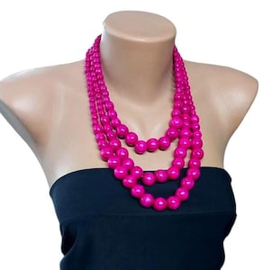 May include: A multi-strand necklace with large, spherical beads in a striking fuchsia hue. The necklace is presented on a mannequin, complemented by a black strapless top. The beads are evenly spaced, displaying a glossy appearance. A statement piece.
