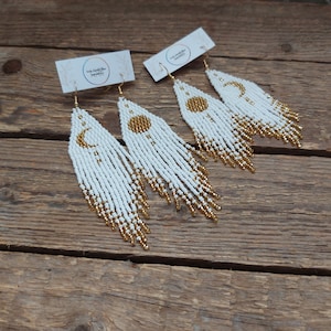 Op de afbeelding: Drie paar witte en gouden kralen oorbellen met een zon en maan ontwerp. De oorbellen zijn lang en hangen.