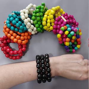 May include: A set of colorful wooden bead bracelets in various colors, including red, orange, yellow, green, blue, and purple. The bracelets are stacked on top of each other. A separate black wooden bead bracelet is shown on a person's wrist.