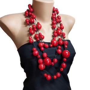 May include: A statement necklace with multiple strands of vibrant red beads. The beads are of varying sizes and are strung on black cord. The necklace is displayed on a mannequin, highlighting its design. The beads are approximately 1-3 cm in diameter.