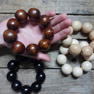 Può includere: Tre bracciali circolari realizzati con grandi perle sferiche lisce. Un bracciale presenta perle marrone scuro lucide, un altro è composto da perle in legno naturale beige chiaro e il terzo è formato da perle nere solide e lucide.