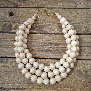 May include: A multi-strand statement necklace featuring three rows of smooth, round, light beige wooden beads. The necklace is finished with a gold-toned metal chain extender and a lobster claw clasp, creating a layered, circular design.