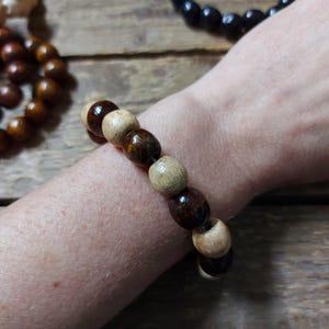 May include: A wooden bead bracelet featuring a mix of light and dark brown beads. The bracelet is worn on a wrist, with other wooden bead bracelets visible in the background. The beads are round and appear to be made of natural wood.