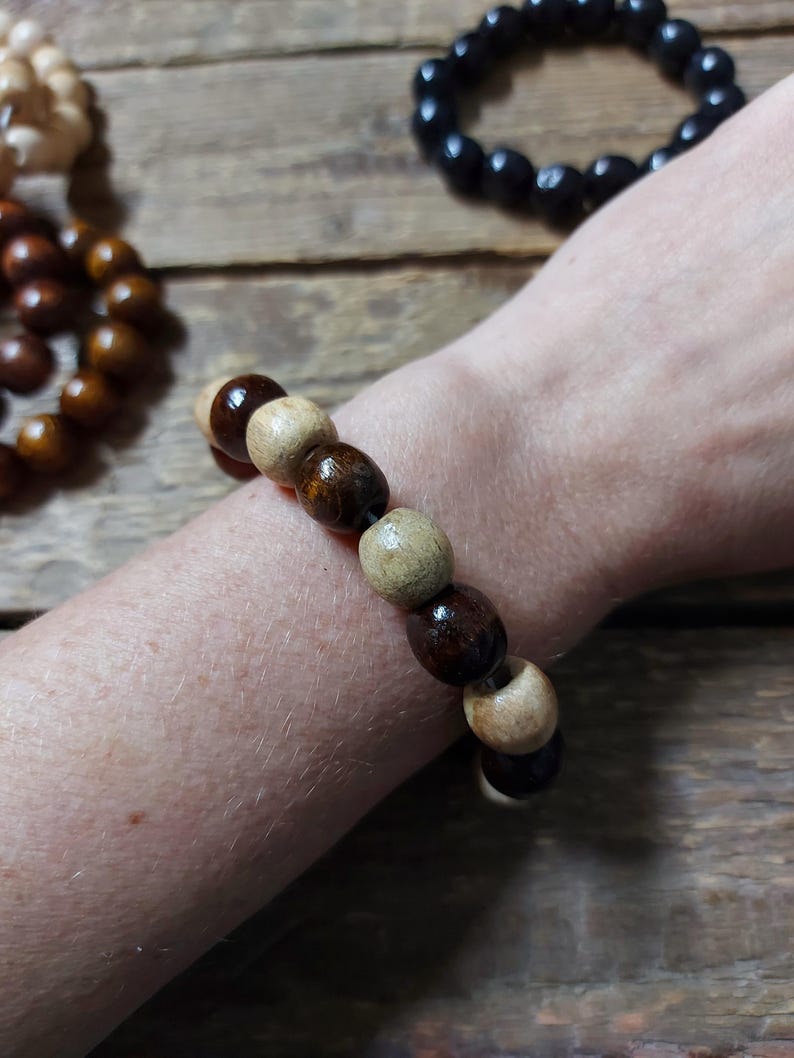May include: A wooden bead bracelet featuring a mix of light and dark brown beads. The bracelet is worn on a wrist, with other wooden bead bracelets visible in the background. The beads are round and appear to be made of natural wood.