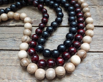 May include: Three strands of wooden beads in natural, dark red, and black colors. The beads are arranged in a circle and are on a wooden surface.