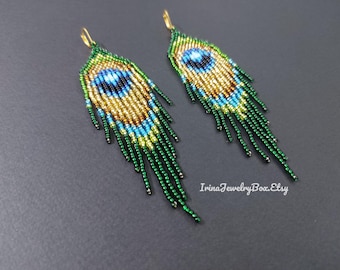 May include: A pair of beaded earrings in a peacock feather design. The earrings feature a teardrop shape with a gold, blue, and green color scheme. The earrings have a fringe of green beads. The earrings are on a dark gray background.