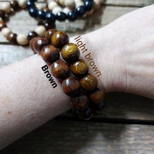 May include: Wooden bead bracelets in various colors, including brown, light brown, black, and cream. The bracelets are shown stacked on a wrist, with the beads appearing to be approximately 0.5 inches in diameter. The background is a wooden surface.