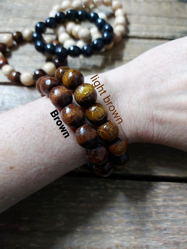 May include: Wooden bead bracelets in various colors, including brown, light brown, black, and cream. The bracelets are shown stacked on a wrist, with the beads appearing to be approximately 0.5 inches in diameter. The background is a wooden surface.