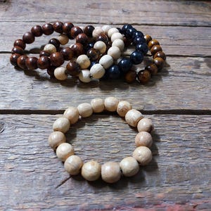 May include: Several wooden bead bracelets in various colors, including light brown, dark brown, white, and navy blue. One bracelet is made of light-colored wooden beads. The bracelets are displayed on a rustic wooden surface.