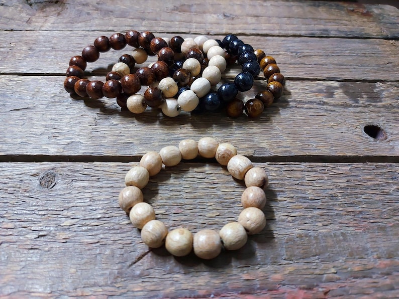 May include: Several wooden bead bracelets in various colors, including light brown, dark brown, white, and navy blue. One bracelet is made of light-colored wooden beads. The bracelets are displayed on a rustic wooden surface.