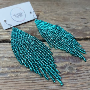 May include: Teal blue beaded dangle earrings with silver hooks. The earrings are made of small beads that are strung together to create a long, flowing design.