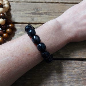 May include: A black beaded bracelet with oval-shaped beads. The bracelet is worn on a wrist, with a wooden surface in the background. Another bracelet with light brown and dark brown beads is partially visible.