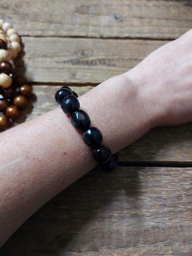 May include: A black beaded bracelet with oval-shaped beads. The bracelet is worn on a wrist, with a wooden surface in the background. Another bracelet with light brown and dark brown beads is partially visible.