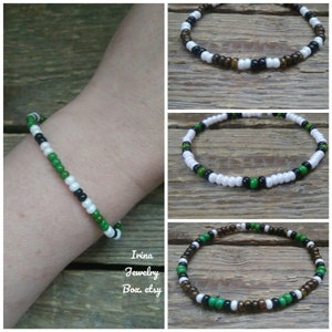 以下が含まれることがあります： ビーズブレスレットのクローズアップ画像が3枚。最初のブレスレットは黒、白、緑。2つ目のブレスレットは白、黒、緑。3つ目のブレスレットは茶色、白、黒、緑。ブレスレットはすべて木製の表面に置かれています。