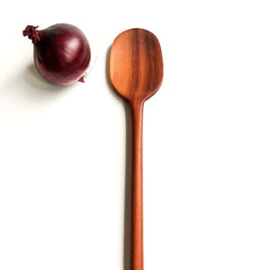 Op de afbeelding: Een houten lepel met een lange, slanke steel en een afgerond, ovaalvormig kop. De lepel is gemaakt van roodbruin hout en heeft een natuurlijke, onbewerkte afwerking.