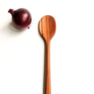 Op de afbeelding: Een lange, houten lepel met een afgerond, ovaalvormig kopje. De lepel is gemaakt van lichtbruin hout met donkerbruine strepen.