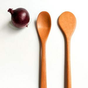 Op de afbeelding: Twee houten lepels in natuurlijke bruine kleur, een met een afgerond uiteinde en de andere met een licht puntig uiteinde. De lepels liggen op een witte achtergrond.
