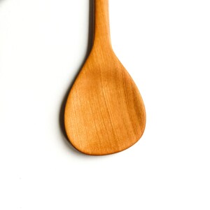 May include: A wooden spoon with a light brown finish. The spoon has a rounded, teardrop-shaped head and a long, slender handle.