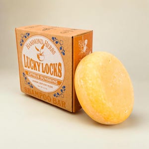 May include: A brown cardboard box with the text "Hammond Herbs Lucky Locks Citrus Sunshine Shampoo Bar" printed on it.  Next to the box is a round, yellow shampoo bar.