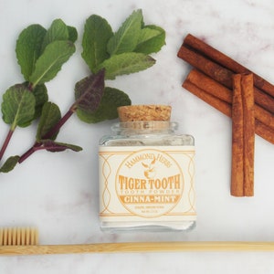 May include: A glass jar with a cork lid filled with white powder. The label reads "Hammond Herbs Tiger Tooth Tooth Powder Cinna-Mint".  A sprig of green mint leaves, cinnamon sticks, and a bamboo toothbrush are also pictured.